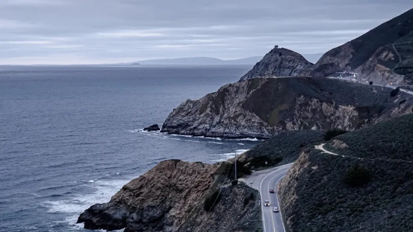 A coastal highway cutting along steep cliffs above the ocean at dusk, the curve of the road lit by passing headlights.