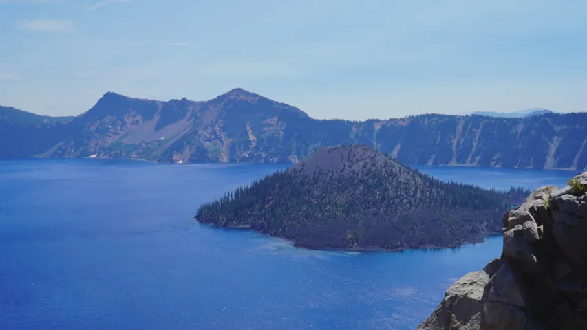 Crater Lake seen from above, deep blue water surrounding the forested cone of Wizard Island, with the caldera rim beyond.
