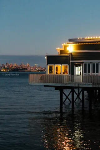 A dark wooden building on stilts over calm water at dusk, its windows lit warm, a city skyline small on the horizon behind it.