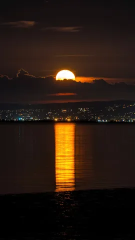 The full moon rising through a cloud bank over the far shore of the bay, the town's lights strung beneath it and a long orange reflection reaching all the way across the water.