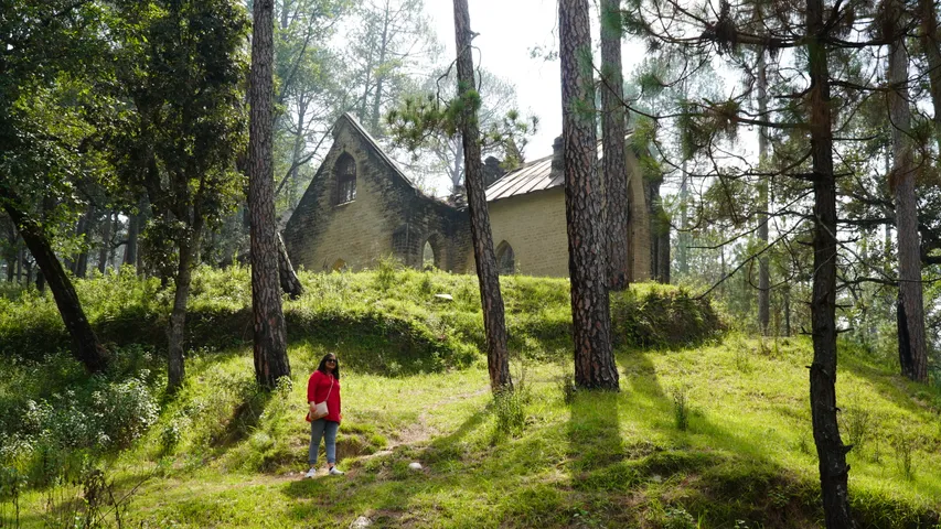 A British era stone and wood church standing among tall pines on a green slope in Ranikhet, Uttarakhand, sunlight coming through the trees, one small figure in red on the path below it.