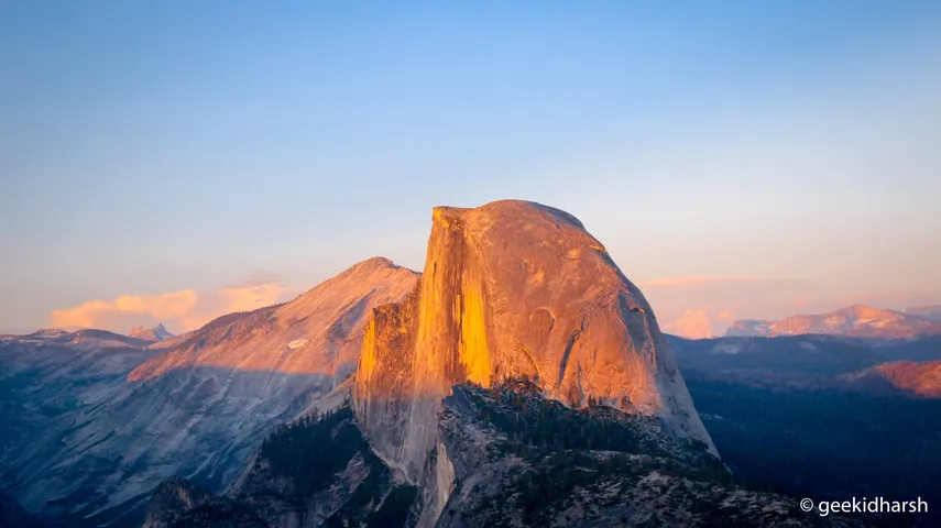 Half Dome in Yosemite at last light, the granite face lit orange down its vertical seam while the valley below has gone into shadow, and the Sierra ridgeline still pink in the distance.