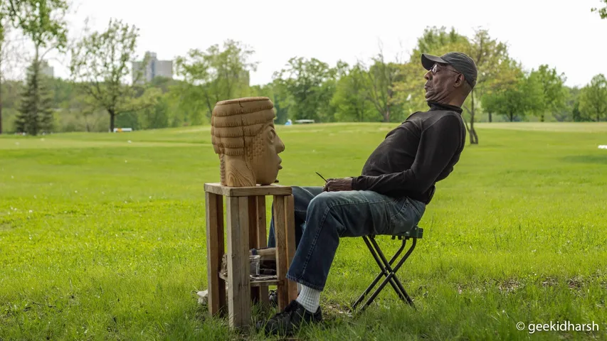 A man in a cap sits on a folding chair on a wide lawn in Forest Park beside a carved wooden head on a pedestal, trees and the city skyline far behind.