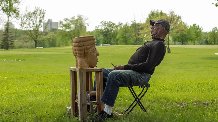 A man in a cap sits on a folding chair on a lawn in Forest Park, St. Louis, beside a carved wooden head on a stand