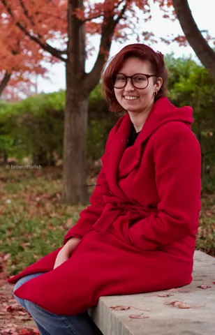A woman in glasses and a long red coat sits on the ground under autumn trees in Forest Park, St. Louis, smiling