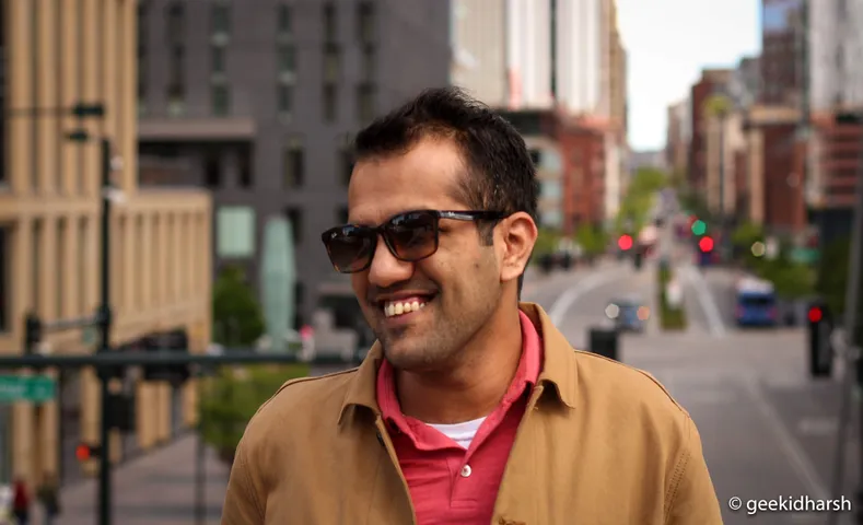 Man in sunglasses and a tan jacket laughing on a downtown street, traffic blurred behind