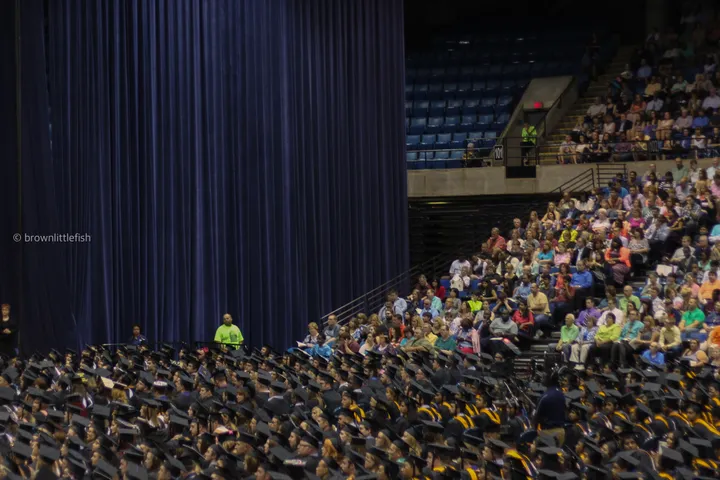 A University of Illinois graduation crowd in caps and gowns, seen from high in the stands, rows of seats and a stage in the distance