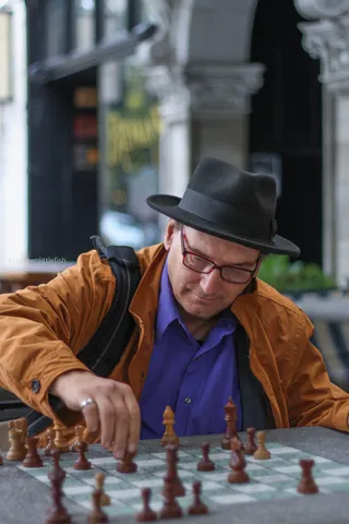 A man in a fedora, glasses and a tan jacket leans over a chessboard on a sidewalk in the Central West End, St. Louis, moving a piece