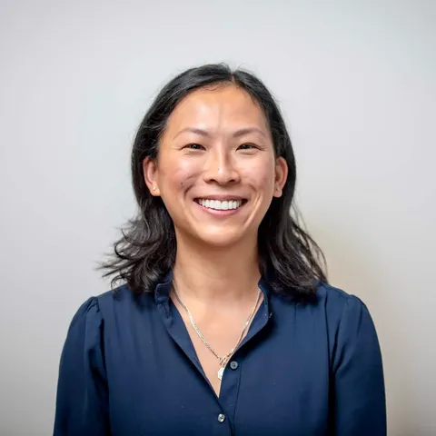 A woman with shoulder-length dark hair in a navy blouse laughing broadly at the camera against a plain warm grey backdrop.