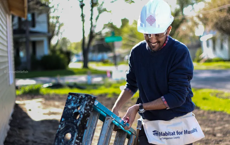 A man in a white cap and dark sweater smiles while working at a wooden frame outdoors in Illinois, a Habitat for Humanity vest on