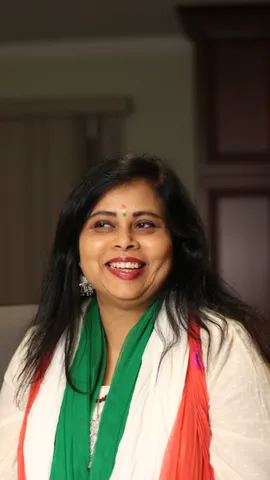 A woman with long dark hair in a white kurta and a green, white and orange dupatta laughs looking to her left, indoors in warm light, dark cabinets soft behind her.