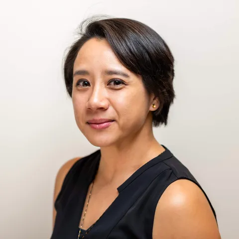A woman with short dark hair in a sleeveless black top, head tilted slightly, looking at the camera with a small smile against a plain light backdrop.