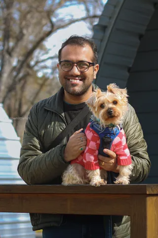 A man in glasses and a green jacket holds a small terrier in a pink and red sweater in the Yosemite snow, both facing the camera