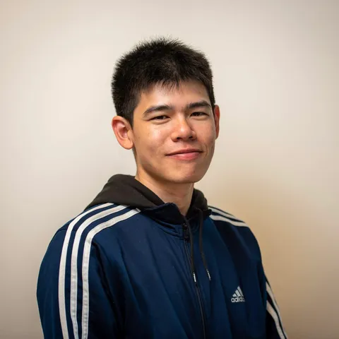 A young man in a navy track jacket with white stripes smiling slightly at the camera against a plain warm grey backdrop.