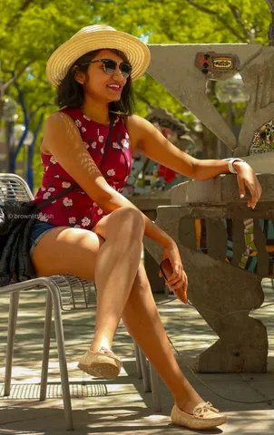 A woman in a straw hat and a red floral top sits on a park bench in Denver with her legs crossed, smiling, trees behind