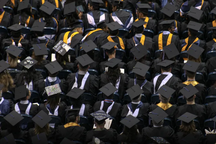 A crowd of graduates seen from behind, black caps with gold tassels filling the frame