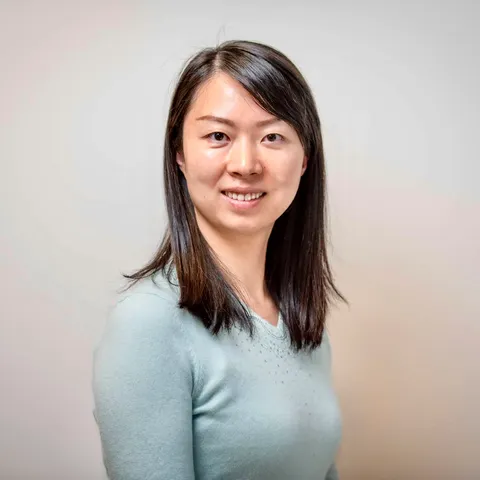 A woman with long dark hair in a pale blue sweater smiling softly at the camera against a plain warm grey backdrop.
