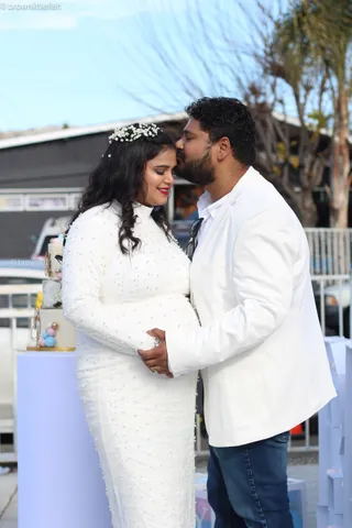 A couple in white at a gender reveal in San Jose, she in a beaded dress with a flower in her hair, he kisses her forehead