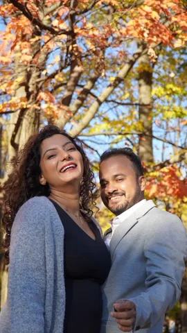 A couple under orange and yellow fall leaves, she laughing in a grey cardigan, he smiling beside her in a light grey blazer.