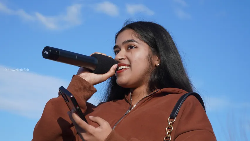 A young woman in a brown jacket sings into a handheld microphone outdoors in Fremont, a bright sky behind