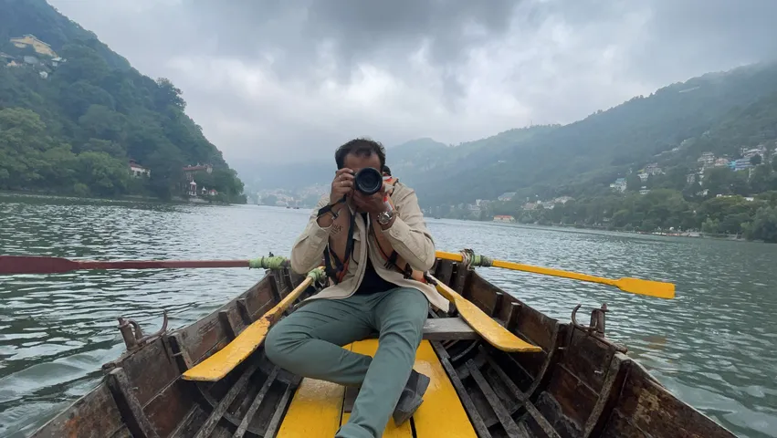 A person seated in the bow of a wooden rowboat holding a camera to their face, yellow oars out, misty green hills and low cloud over the lake behind.