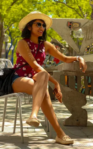 Woman in a straw hat and floral top sitting on a park bench, smiling, trees behind