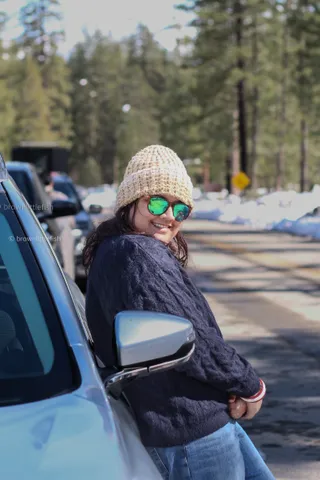 A woman in a cream knit hat and mirrored sunglasses leans out of a car window on a snowy road in Yosemite, smiling, pines behind