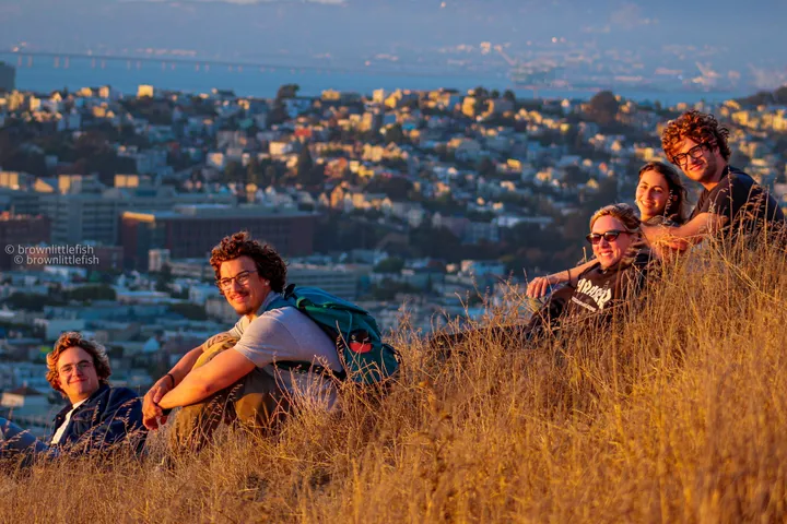 Five friends sitting in tall dry grass on a hillside at sunset above San Francisco, the city and the bay below them