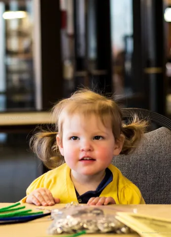 A toddler in a yellow top with two small pigtails looks straight at the camera across a breakfast table in Illinois
