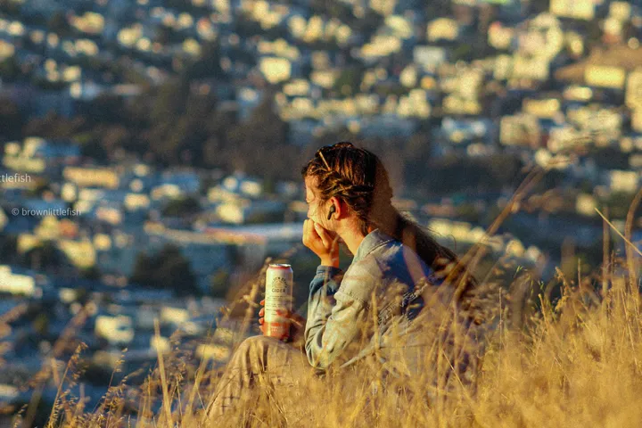 A man seen from behind in tall dry grass on a hillside at sunset, a can in his hand, San Francisco below