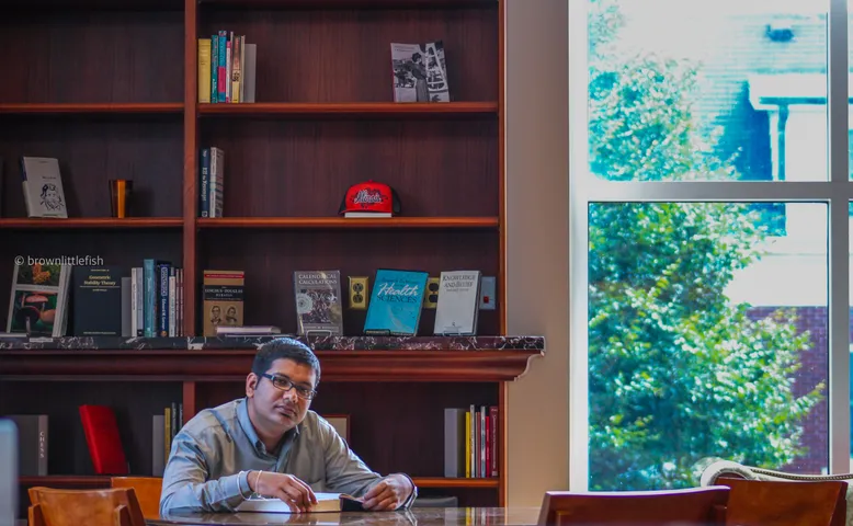 A man in glasses seated at a wooden desk in an office in Champaign, Illinois, bookshelves and framed pictures behind him