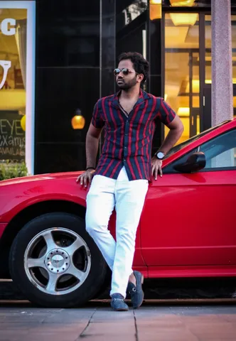A man in sunglasses and a red and navy striped shirt leaning against the front of a red car outside a lit storefront, one hand on his hip.