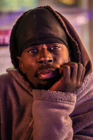 A close portrait of a man in a dark hoodie with the hood up, hand at his chin, lit by city light, St. Louis