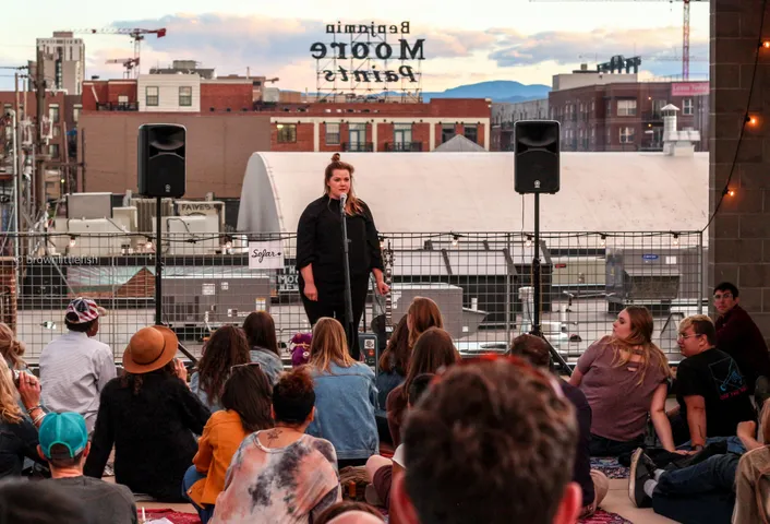 A singer on a rooftop stage at dusk in Denver, speakers either side, the backs of a seated crowd in the foreground