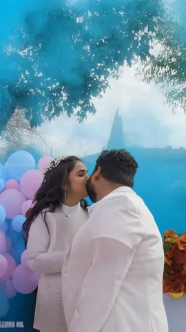A couple in white kiss under trees at a gender reveal in San Jose, pink and blue balloons and coloured smoke behind them
