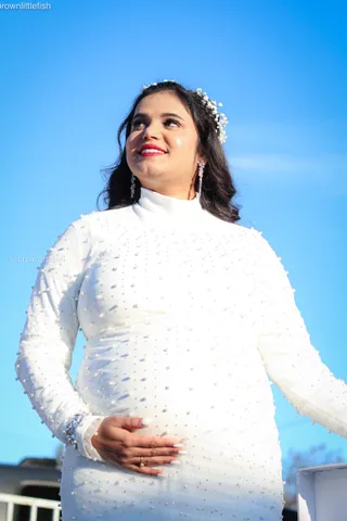 A woman in a white beaded dress with a flower in her hair holds her pregnant belly, looking up at a clear blue sky