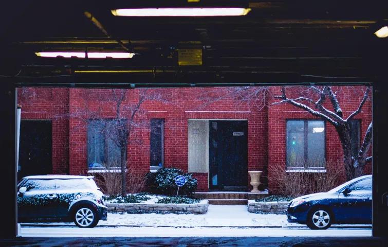 A red brick row house at night, its stoop and two parked cars under fresh snow, a bare tree in front.