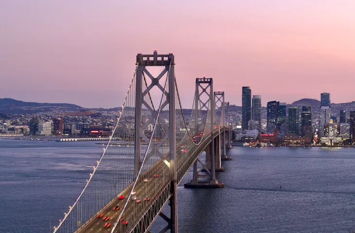 The Bay Bridge at sunset, its towers lit against a pink sky, the San Francisco skyline behind it and the bay below.