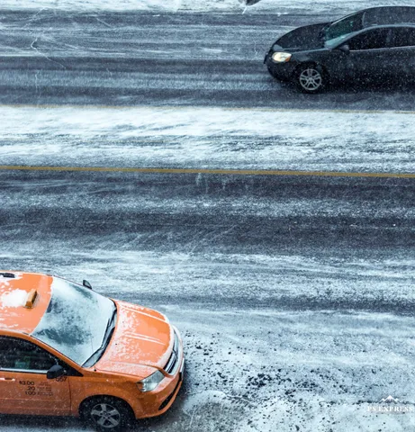 Seen from above, an orange taxi covered in snow drives along a snow-packed street, tyre tracks in the white, a dark car ahead.