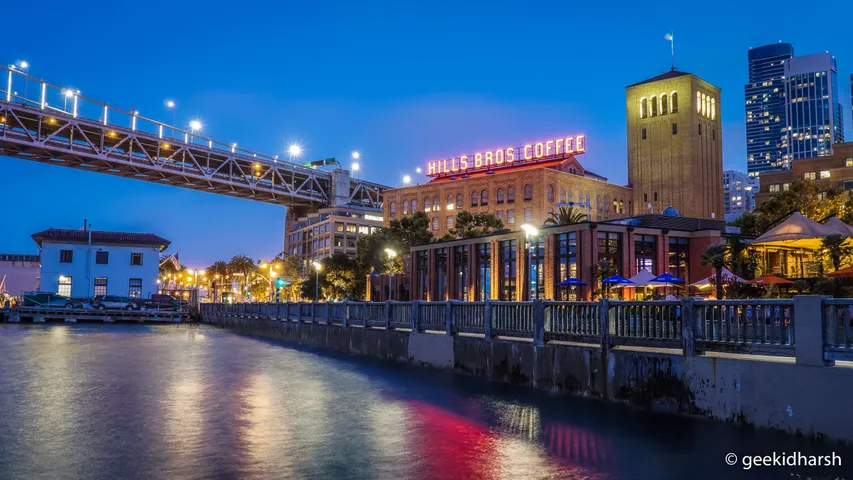 The Hills Bros Coffee sign glowing red on its brick building at the foot of the Bay Bridge in San Francisco, the bridge deck crossing the top of the frame and the sign's reflection running down the water along the Embarcadero.