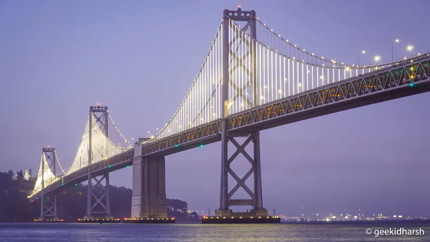 The western span of the Bay Bridge at dusk from the San Francisco waterfront, its cables lit white against a lavender sky, Yerba Buena Island low on the left and the piers picked out in small lights below.