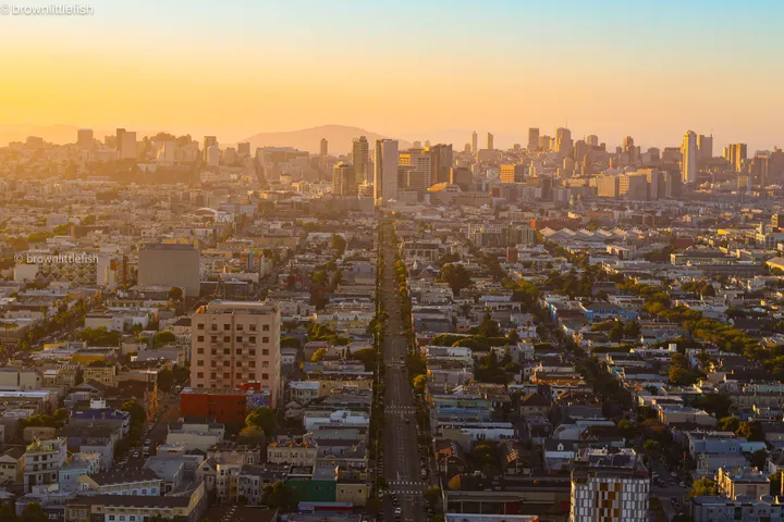 San Francisco seen from a hill at golden hour, a long straight avenue running to the downtown towers, haze over the bay