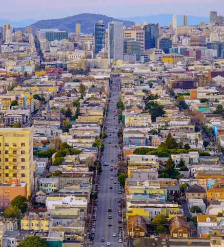 Aerial view down a long straight street lined with pastel houses, running toward the downtown San Francisco skyline under hazy afternoon light.