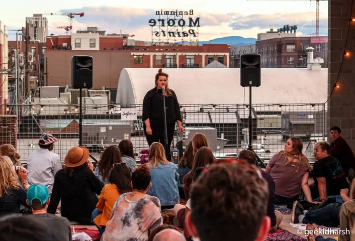 Crowd seated on a rooftop in Denver watching a performer at dusk, city buildings behind