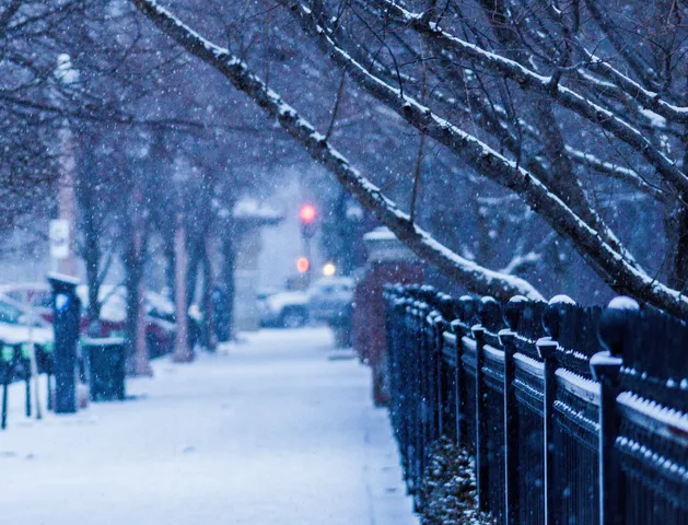 Snow falling on an empty sidewalk, a wrought iron fence running away to the right beneath bare branches, parked cars and a red traffic light going soft in the distance.