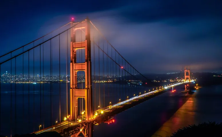 The Golden Gate Bridge at night seen from the Marin headlands, fog spilling over the north tower, headlights drawn into one continuous white line along the deck, and the lights of San Francisco across the water behind it.