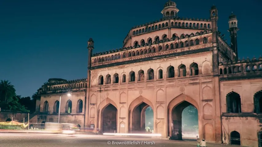 Rumi Darwaza, the Mughal-era gate at the Bara Imambara complex in Lucknow, lit from below at night. Three tiers of arches and carved parapets rise against a deep blue sky, with a vehicle's lights drawn into a long streak across the road at its foot.