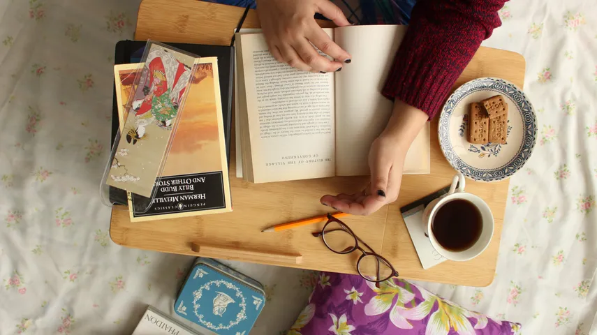 Looking straight down at a wooden lap tray on a floral bedspread: two hands holding a paperback open, a stack of books and an illustrated postcard beside them, a cup of black coffee, shortbread on a blue and white plate, and a pair of glasses set down on the wood.
